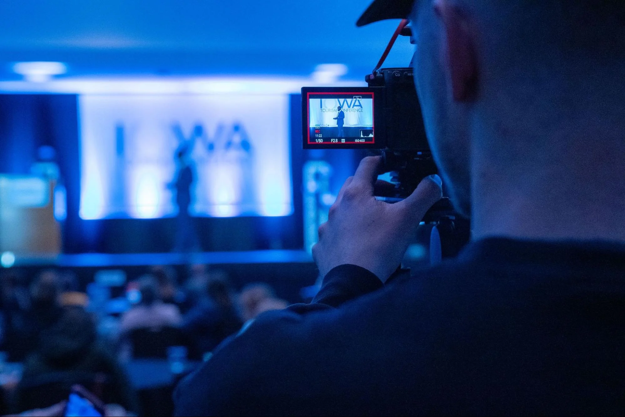 Videographer filming a live event with a camera as speakers appear on stage in the background